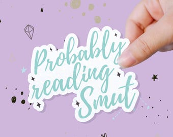 May include: A white sticker with the text "Probably reading Smut" in a teal script font. The sticker is held by a hand against a light purple background with scattered star and diamond shapes.