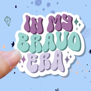 May include: A sticker with the text "In My Bravo Era" in a retro, colorful font. The sticker has a white border and is surrounded by a light blue background with stars and other doodles.