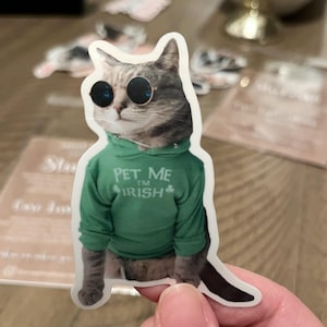 May include: A sticker featuring a cat wearing sunglasses and a green hoodie. The hoodie has the text "PET ME I'M IRISH" with a shamrock. The cat has gray and brown fur and is wearing round, black sunglasses.