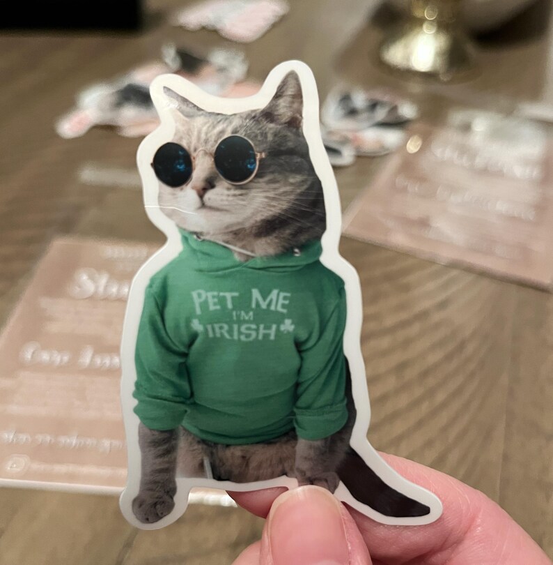 May include: A sticker featuring a cat wearing sunglasses and a green hoodie. The hoodie has the text "PET ME I'M IRISH" with a shamrock. The cat has gray and brown fur and is wearing round, black sunglasses.