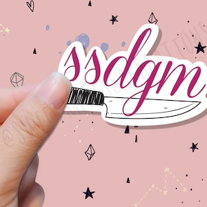 Peut inclure: Un autocollant rose et blanc avec un dessin noir et blanc d'un couteau. L'autocollant porte le texte "ssdgm" écrit en cursive rose.