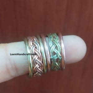 Pode incluir: Um conjunto de anéis empilhados com uma mistura de tons de cobre e latão. O design inclui duas bandas trançadas e duas bandas lisas e simples. O texto LaxmiHandicraftitems é visível à esquerda. O metal tem um acabamento polido.