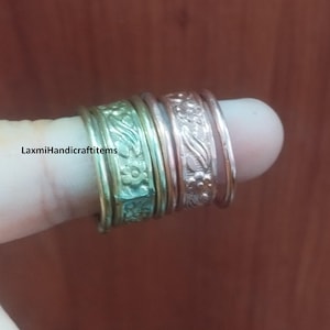 Pode incluir: Um conjunto de anéis empilhados com uma banda de tom dourado com um padrão floral em relevo e uma banda de tom cobre com um design floral correspondente. O texto LaxmiHandicraftitems é visível no lado esquerdo da banda dourada.