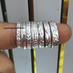 May include: A set of seven silver-toned metal rings with various embossed patterns including leaf motifs, geometric shapes, and floral swirls, featuring the text LaxmiHandicraftitems printed on the left side of the image.