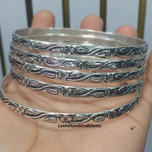 Könnte beinhalten: Ein Set aus fünf silberfarbenen Metallarmreifen mit einem durchgehenden geprägten Blumen- und Rankenmuster. Die Oberfläche jedes Armreifs ist poliert und weist abgedunkelte vertiefte Bereiche auf, um das komplexe botanische Design hervorzuheben. Der Text LaxmiHandicraftitems ist schwarz auf dem untersten Armreif aufgedruckt.