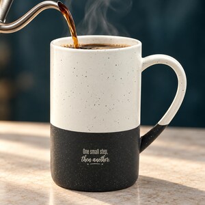 Peut inclure: Un grand mug en céramique bicolore avec une finition mouchetée. La moitié supérieure est blanc cassé et la moitié inférieure est noire. La partie noire porte l'inscription blanche "One small step, then another". Du café chaud est versé.
