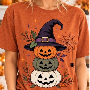 Puede incluir: Una camiseta de manga corta color naranja quemado con un estampado gráfico de tres calabazas apiladas. La calabaza superior lleva un sombrero de bruja morado. El diseño incluye una telaraña, una pequeña araña y hojas de otoño con estrellas.
