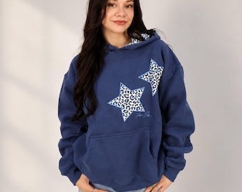 May include: A navy blue hooded sweatshirt featuring two large star-shaped appliques with a white and blue leopard print pattern. The hoodie includes a front kangaroo pocket and a matching leopard print lining inside the hood.