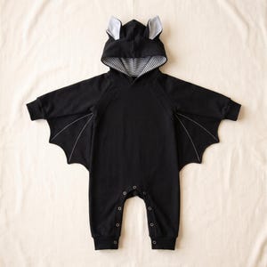 May include: A black long-sleeve hooded romper featuring bat-wing extensions under the arms with white stitched detailing. The hood includes two small ears with light gray inner accents and a black and white striped interior lining.