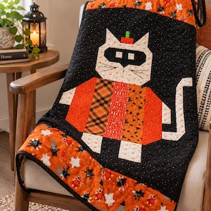 May include: A quilted throw blanket featuring a central cat figure wearing a black mask and a small orange hat. The cat is composed of various orange and black patterned fabrics. The background is black with white speckles and orange borders.