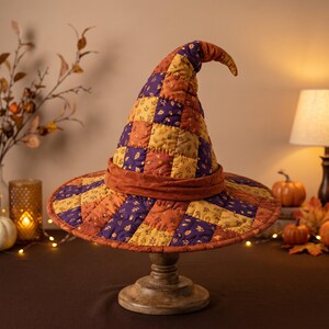 May include: A quilted witch hat featuring a patchwork design of squares in shades of purple, orange, and yellow, decorated with a repeating pattern of small leaves and acorns, finished with a solid orange band around the base.