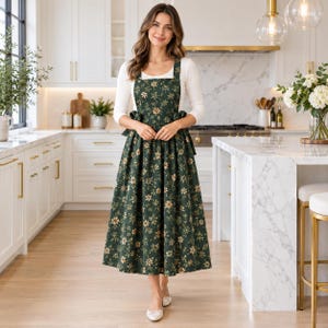 May include: A dark green pinafore apron dress featuring a repeating pattern of small white and yellow flowers. The garment has a square neckline, wide shoulder straps, and a full, pleated skirt that falls to the mid-calf, worn over a white long-sleeved top.