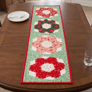 Puede incluir: Un camino de mesa acolchado rectangular con cuatro grandes motivos octogonales en patrones florales alternos de color rojo, verde y rosa. El camino tiene un fondo verde claro con un pequeño estampado de hojas y un borde rojo brillante.
