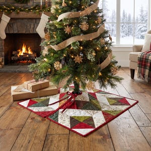 Op de afbeelding: Een vierkante kerstboomkleed van stof met een patchworkontwerp van rode, donkergroene, bruine en crèmekleurige stoffen vierkanten met ingewikkelde patronen. Het kleed heeft een rode rand en rode stoffen linten bij de opening in het midden.