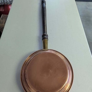 May include: A round copper bed warmer featuring a long, dark brown wooden handle with a brass ferrule connecting the handle to the copper pan. The pan has a smooth, metallic surface and a small brass loop attached to the bottom edge for hanging.