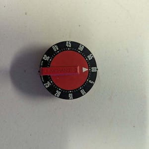 May include: A round mechanical kitchen timer with a black outer ring featuring white numbers from 5 to 60. The centre is a red circular dial with a red horizontal arm labelled JUNGHANS and a white triangular indicator pointing towards the numbers.