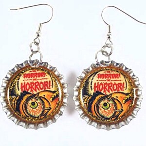 May include: A pair of silver earrings with bottle caps featuring a vintage horror movie poster with a large eye and the text "The Horror! The Horror!" printed in red.