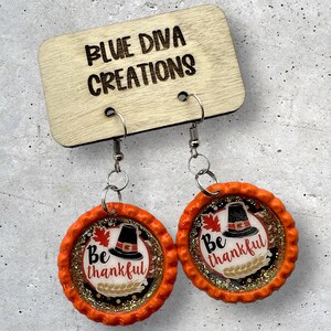 May include: A pair of orange and gold earrings with a pilgrim hat and the words "Be thankful" on each earring.