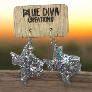 May include: Silver glitter Texas-shaped earrings hanging from a wooden display with the text "BLUE DIVA CREATIONS". The earrings are suspended by silver hooks. The background has a blurred sunset.