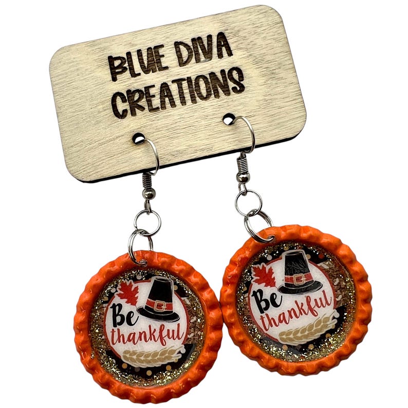 May include: A pair of orange and gold glitter earrings with a pilgrim hat and the text "Be thankful" on each earring.