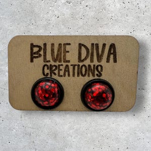 May include: A pair of red glitter stud earrings with black backing. The earrings are displayed on a wooden card with the text "Blue Diva Creations".