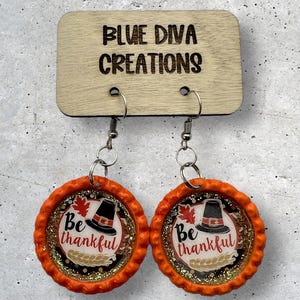 May include: A pair of orange and gold Thanksgiving themed earrings. The earrings are made from bottle caps and feature a pilgrim hat, leaves, and the text "Be thankful".