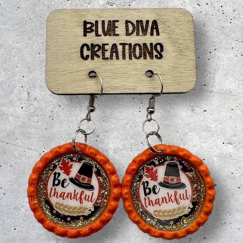 May include: A pair of orange and gold Thanksgiving themed earrings. The earrings are made from bottle caps and feature a pilgrim hat, leaves, and the text "Be thankful".