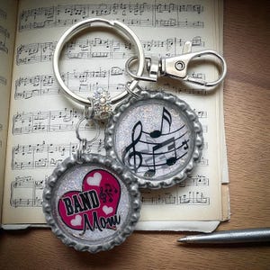 May include: A silver keychain with two bottle cap charms. One charm has musical notes on a glitter background. The other charm has a pink heart with the words "BAND Mom" and musical notes. The keychain is on a sheet of music.