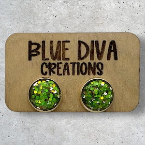 May include: A pair of green glitter stud earrings with gold settings. The earrings are displayed on a wooden card with the text "Blue Diva Creations".