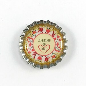 Peut inclure: Un bouchon de bouteille doré avec un fond blanc et un motif floral rose. Le texte "LIFETIME OF LOVE" est imprimé en noir avec un cœur rouge.