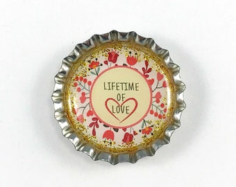 May include: A gold bottle cap with a white background and a pink floral design. The text "LIFETIME OF LOVE" is printed in black with a red heart.