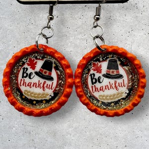 May include: A pair of orange and gold glitter Thanksgiving themed earrings. Each earring features a pilgrim hat, a leaf, and the text "Be thankful".