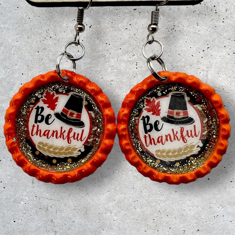 May include: A pair of orange and gold glitter Thanksgiving themed earrings. Each earring features a pilgrim hat, a leaf, and the text "Be thankful".