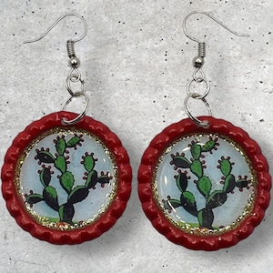 May include: A pair of red and gold earrings featuring a cactus design. The earrings are made from bottle caps and have silver hooks.