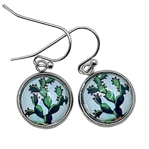 May include: A pair of silver dangle earrings with a round glass cabochon featuring a green cactus illustration on a light blue background.