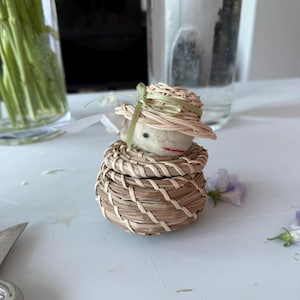 May include: A small, round, woven wicker basket with a matching lid. Resting on top of the lid is a needle-felted figure with a pale, off-white head, a single black eye, a red stitched mouth, and a light tan woven hat tied with a green ribbon.