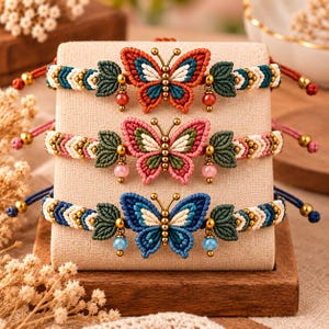 May include: Three macrame bracelets featuring central butterfly motifs in orange, pink, and blue. Each bracelet has braided bands with chevron patterns, small green leaf-shaped accents, and dangling beads in matching colors with gold-tone metal details.