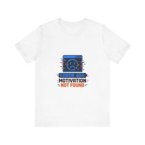 May include: A white short-sleeved t-shirt featuring a pixelated graphic of a blue computer window with a sad face icon. Below the window, the text reads ERROR 404: MOTIVATION NOT FOUND in black and orange, accented by small red and blue glitch lines.