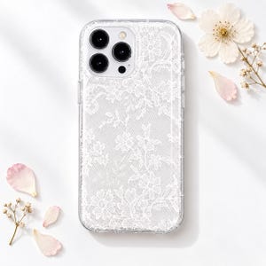 May include: A clear smartphone case featuring a delicate white lace pattern with intricate floral details. The case has a rectangular shape with rounded corners and a cutout for a triple-lens camera system on the upper left side of the device.