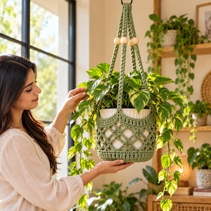 May include: A sage green crocheted plant hanger featuring a lattice pattern and three light-coloured wooden beads at the top. The hanger holds a white ceramic pot containing a trailing pothos plant with variegated green and yellow leaves.