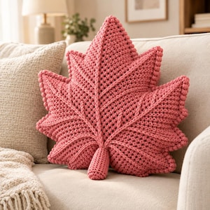 May include: A dusty rose colored throw pillow shaped like a maple leaf. The pillow is crafted with a textured crochet knit pattern, featuring raised central veins and a small stem at the base. The edges are finished with a decorative bobble trim.