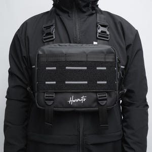Puede incluir: Una bolsa de pecho negra con un compartimento principal rectangular, un panel frontal con correas MOLLE, dos correas verticales ajustables con hebillas y el texto blanco Harmits impreso en la parte inferior frontal, sobre una chaqueta negra.