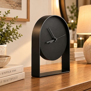 May include: A matte black table clock featuring an arched frame and a circular face. The clock has three thin black hands positioned in the center of the solid black dial, with no numbers or markings on the face of the timepiece.