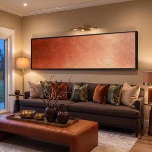 Könnte beinhalten: Ein großes, rechteckiges, horizontales Wandbild mit einer texturierten Farbverlaufsoberfläche in gebrannten Orange- und Kupfertönen, das in einem dünnen schwarzen Rahmen über einem dunkelgrauen Sofa mit verschiedenen Zierkissen hängt.