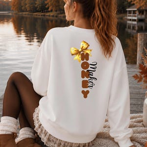 Könnte beinhalten: Ein weißes Langarm-Sweatshirt mit einem Grafikdruck auf der Rückseite. Das Design zeigt eine gelbe Schleife oben, darunter das Wort HONEY in vertikal gestapelten braunen Buchstaben und das Wort Maker in schwarzer Schreibschrift.