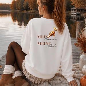 Könnte beinhalten: Ein weißes Langarm-Sweatshirt mit einer Grafik aus einem Honiglöffel und einer Biene sowie dem Text "MEIN Bienenvolk MEINE Regeln", der in brauner und schwarzer Schrift auf der Rückseite des Kleidungsstücks gedruckt ist.