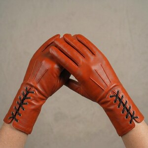May include: A pair of burnt orange leather gloves featuring three vertical stitched lines on the back of the hand and a decorative black lace-up detail with silver-toned metal eyelets at the wrist, extending from the cuff toward the palm.