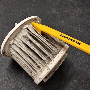 May include: A cylindrical white vacuum cleaner filter covered in grey dust and debris, being cleaned by a yellow plastic tool with the word FARMFIX printed in black letters on its side. The filter has vertical pleats and a circular top rim.
