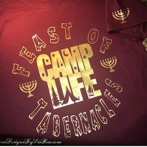 Może przedstawiać: Bordowa koszulka z złotymi literami i grafiką. Projekt zawiera słowa "FEAST OF CAMP LIFE TABERNACLE" w okrągłym układzie, wraz z menorami, koroną, namiotem i innymi symbolami. Na rękawie znajduje się również grafika menory.