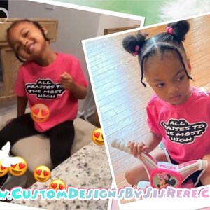 May include: A young child wearing a pink t-shirt with the text "ALL PRAISES TO THE MOST HIGH" in a black and white design. The child is sitting on a chair and smiling. The shirt is paired with black leggings.